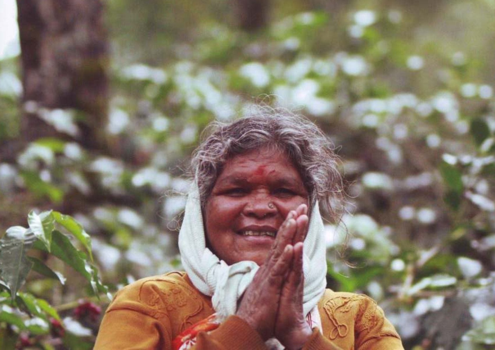 A woman on the BR Hills estate greeting the camera with folded hands, coffee bushes behind her
